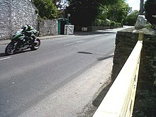 Racing motorcycle rider on left adjacent to a high wall approaching camera position with road stretching out behind to bend in distance