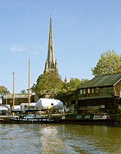 Een hoge kerktoren boven een kade met houten schuren en boten bedekt met dekzeilen.  Daarvoor op het water een tweemaster zeilboot en een narrowboat