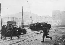 Soldats allemands courant en direction d'un véhicule blindée dans une rue jonchée de débris