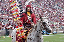 Um cavalo manchado de marrom e branco montado por um mascote dos esportes em trajes indígenas modernos, acenando uma bandeira, fica em um campo de esportes.  Mais pessoas estão visíveis no campo e uma grande multidão enche o estádio ao fundo.