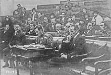 Photographie en noir et blanc d'un homme à l'air soucieux assis dans une sorte d'hémicycle.