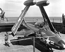 Fotografia em preto e branco de um Banshee com as asas dobradas no elevador de um porta-aviões.