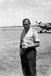 The head and shoulders of a young man, shown in semi-profile. He wears a shirt with an Iron Cross displayed at the front of his shirt collar. His hair is dark and short, his nose is long and straight, and his facial expression is showing a broad smile; gazing at a point to the right of the camera.