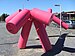Alter Hund, eine rosa Skulptur, die einem Ballontier ähnelt und sein hinteres Bein anhebt, um auf ein Straßenschild mit der Aufschrift "Mein Weg" zu urinieren.