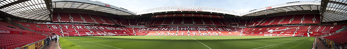 A stand of a football stadium. The seats are red, and the words "Manchester United" are written in white seats. The roof of the stand is supported by a cantilever structure. On the lip of the roof, it reads "Old Trafford Manchester".