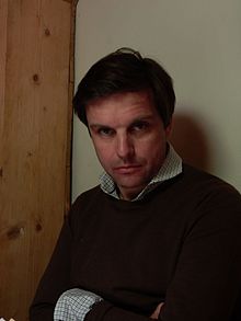 Caucasian man with brown hair, blue eyes, wearing brown sweater over white checked open-collar shirt