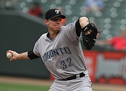 Een man in een grijze baseballtrui en zwarte baseballpet gooit een honkbal met zijn rechterhand.  Zijn trui leest "Toronto" en "34" op de voorkant in zwarte blokletters omlijnd in blauw, en hij heeft een zwarte honkbalhandschoen aan zijn linkerhand.