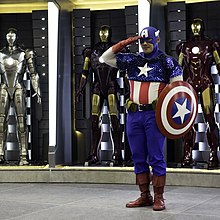 captain America au garde à vous tenant son bouclier de même couleur que son habillement : étoile blanche,plus les couleurs bleu,blanc,rouge,derrière lui des armures