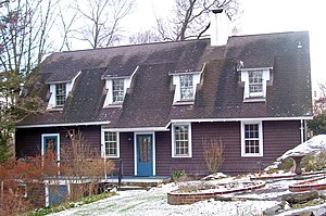 A brown house with low sloping black roof and dormer windows. There is a light snow covering on the brick and stone fountain and front lawn.