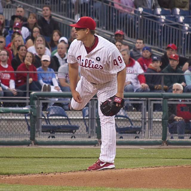 A young man wearing a white baseball uniform pinstriped with red and a red baseball cap follows through after throwing an unseen baseball