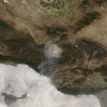 A satellite picture of the fire from directly overhead shows a round, brown cloud of smoke with a white pyrocumulus cloud bubbling in its center, surrounded by rugged mountains