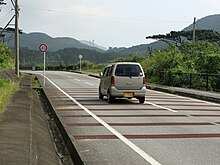 Photo couleur : une section de route traversée perpendiculairement de zébras. Un paysage montagneux en arrière-plan.