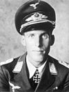 The head of a young man, shown in semi-profile. He wears a peaked cap, a military uniform with a military decoration in shape of an iron cross displayed at the front of his shirt collar.