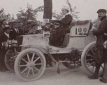 Foto di un'auto il cui aspetto ricorda ancora una carrozza trainata da cavalli, guidata da una donna.