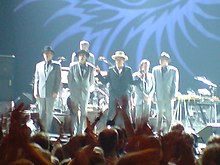 heads of people cheering in front of Dylan acknowledging five band members for the most part in grey suits wearing black hats