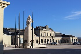 Edifício horizontal com frente a uma praça e uma estátua no gramado verde.