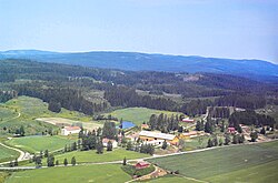 flyfoto av gårdsanlegg med flere bygninger. Dyrket mark i forgrunnen, skog i bakgrunnen