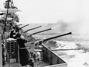 The flank of a ship. Several long-barrelled guns are aimed over the side, and are being operated by sailors. One of the guns has just fired, with a cloud of smoke issuing from the barrel.