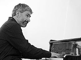 un homme souriant, cheveux gris et barbe, assis au piano