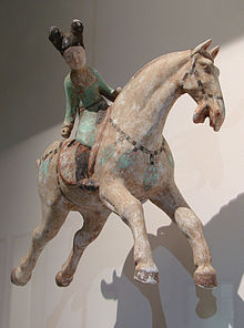 keramická figurka jezdkyně na cválajícím koni, vybledlé barvy: modrozelený přiléhavý oděv ženy, vysoké černé boty, černé vlasy vyčesané do dvou drdolů čnících do výšky, kůň má černou uzdu a řemen s modrozelenými ozdobami, sedlo je hnědé