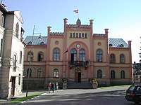 Concello de Kuldiga, un edificio con tellado a dúas augas cunha fachada pintada en vermello e marrón e cunha bandeira de Letonia na parte superior do edificio.
