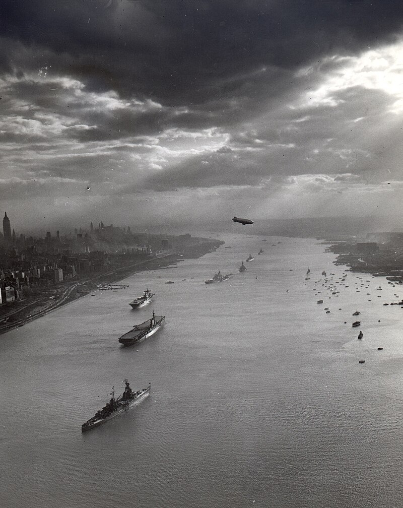 Tugboats and U.S. Navy warships pictured in the Hudson River with the New York City skyline in the background for the Navy Day celebrations on 27 October 1945. Visible in the foreground are the anchored warships USS Augusta (CA-31), USS Midway (CVB-41), USS Enterprise (CV-6), USS Missouri (BB-63), USS New York (BB-34), USS Helena (CA-75), and USS Macon (CA-132)