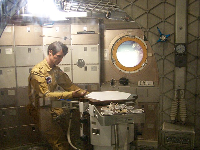 A mockup of the inside of Skylab at the Smithsonian, based on the Skylab B module. In the center, a dummy dressed in a gold jumpsuit sits at a table. Behind him are white cabinets that hold the crews equipment. Off to the right, a porthole shows a view of the Smithsonian.