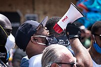 African American man wearing white shouting into bullhorn pointed upper right