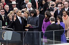 Barack Obama holds his right hand in the air as Chief Justice John Roberts administers the oath of office to him. Michelle Obama looks at him with Malia and Sasha Obama.