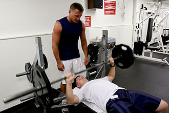 Bench press with a bench and barbell. The man standing is 'spotting' and ready to help with lifting the weight if the lifter becomes too fatigued. Bench press 1.jpg