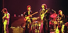 Dylan avec trois musiciens de The Band sur scène. Dylan est le troisième à partir de la gauche, vêtu d'une veste et d'un pantalon noirs. Il chante et joue de la guitare électrique.