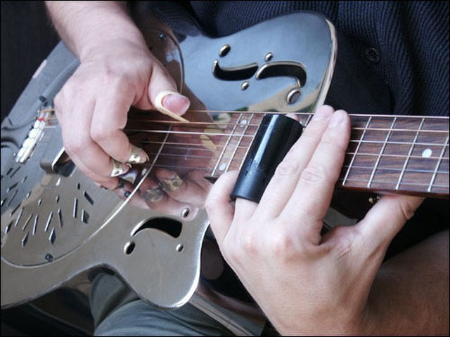 A musician playing slide guitar style; The slide is on his left ring finger. He is playing a metal-body resonator guitar (a National-type) using finge