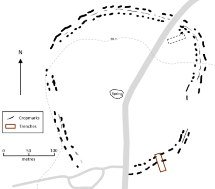 Two concentric more-or-less circular dashed lines, indicating cropmarks, with a thin line occasionally visible between them. A road passes through north to south. A rectangular trench cuts the enclsoure line in the southeast corner and there is a small rectangular shape inside the enclosure in the northeast corner.