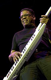 A man wearing glasses, with his eyes closed, playing a white keytar with black and white keys.