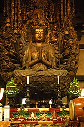 Front view of a cross-legged seated statue with many arms positioned in a circular fashion around the body carrying various objects like bells and staffs. One pair of hands is folded in front of the body in prayer. The statue is positioned on a flower-shaped platform. Color photograph.