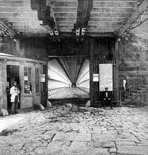 A square opening forms the entrance to the bridge's lower level. Vertical wooden supports line the level. Flagstones pave the entrance and a booth stands to the left. A man stands at the booth, another sits in front of the entrance, and another is walking up the stairs on the right.