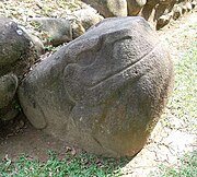 A smoothly finished boulder carved into the shape of a seated frog or toad, with the head raised to the right and sporting a prominent smile. The eyes, mouth, nostrils and legs are all carved in low relief. The sculpture is set against the stonework base of a structure, behind it at left.
