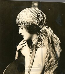 A young white woman with dark hair, photographed in profile, wearing a striped head scarf tied at the nape