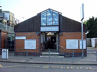 A brown-bricked building with a rectangular, light blue sign reading "WEST HARROW STATION" in white letters all under a light blue sky