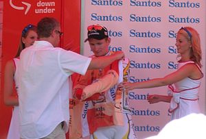 A cyclist is presented an orange jersey, and is in the process of donning it. A man wearing white is facing the cyclist and pulling the jersey's sleeves forward around him. Two women in white dresses flank the cyclist.