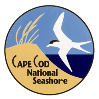 A circular logo depicting a sand dune topped with beach grass next to the ocean, with a white sea bird flying overhead.