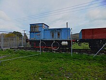 A small blue diesel locomotive, attached to a brown wagon, on a section of track.