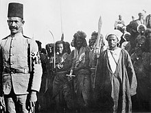 A group of Hadendoa Sudanese prisoners bearing weapons. An Egyptian military policeman stands in the foreground. General Kitchener and the Anglo-egyptian Nile Campaign, 1898 HU93854.jpg