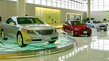 Car showroom displaying three sedans, the nearest on a glass turntable, in front of a reception counter and windows.