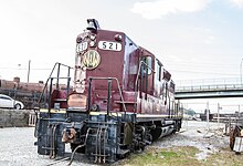 A diesel locomotive idling in the yard with an abandoned steam locomotive and a car in the background