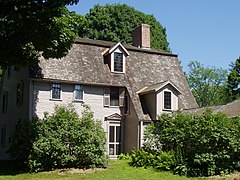 A light brown colonial frame house with wooden roof shingles. The right side of the house front has an addition where the roof extends to the first floor.