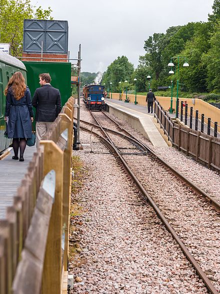 A Bluebell Railway heritage train arrives into the Bluebell's East Grinstead station platform, during the railway's 2013 Edwardian weekend. The two people on the left are walking along the heritage line's Observation platform. To the right, beyond the second fence where the guard is, is located the connecting line to Network Rail