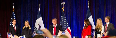 The Trump family at a 2016 campaign event. From left to right are son-in-law Jared, eldest daughter Ivanka, Donald himself, wife Melania, daughter-in-law Lara, and second-eldest son Eric. They are all standing behind a podium with the word "TRUMP" printed in white-on-blue text. Behind them are some American flags hanging on poles, as well as a blue curtain. In the foreground are audience members taking the Trumps' pictures.