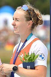 Nika Johanna Vos schaut bei der Siegerehrung nach rechts. Sie trägt das niederländische Nationaltrikot über einem weißen T-Shirt. Um den Hals hängt ein blaues Band, an dem die nicht sichtbare Silbermedaille befestigt ist. Ihre Sonnenbrille hat sie hochgeschoben. In der rechten Armbeuge hat sie einen Blumentopf.