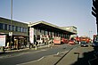 A grey building with windows stands under a clear, blue sky as people mill about on the sidewalk in front of the building and vehicles traverse the foreground.